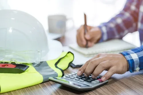 Pessoa a calcular e a tirar notas numa secretária com capacete e colete de segurança, simbolizando o planeamento de obras ou orçamento habitacional.