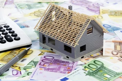 Miniatura de uma casa em construção sobre notas de euro, com uma calculadora e uma caneta ao lado, simbolizando o investimento ou financiamento imobiliário.